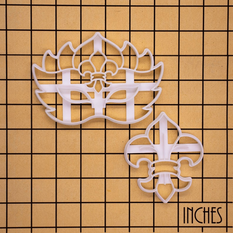 K&ouml;nnte beinhalten: Zwei wei&szlig;e Plastik-Ausstecher, einer in Form einer Fleur-de-lis und der andere in Form einer Maske mit einer Fleur-de-lis in der Mitte. Die Ausstecher befinden sich auf einem braunen Gitterhintergrund mit dem Wort "INCHES" in der unteren rechten Ecke.