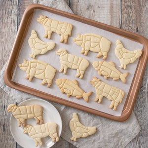 Puede incluir: Galletas horneadas con forma de animales, incluyendo una vaca, un cerdo, un pollo, un pez y una oveja, con l&iacute;neas de corte. Las galletas est&aacute;n dispuestas en una bandeja para hornear y un plato blanco, de color marr&oacute;n claro.