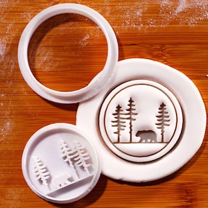 Könnte beinhalten: Weißer Keksausstecher mit einem Wald- und Bärenmotiv. Der Stempel enthält einen runden Ausstecher und zwei Abdrücke des Designs. Das Design zeigt Kiefern und einen Bären, der vor den Bäumen geht.