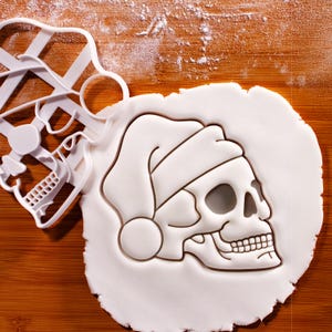 May include: A white skull-shaped cookie cutter with a Santa hat design next to a skull-shaped cookie on a wooden surface dusted with flour. The cookie cutter is made of white plastic and has a detailed skull shape.