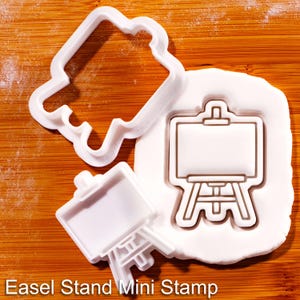 May include: White plastic cookie cutters in the shape of an easel stand and a rectangular shape. The text "Easel Stand Mini Stamp" is printed at the bottom of the image. The cutters are on a wooden surface.