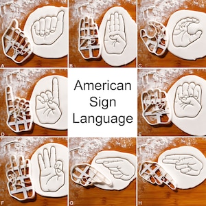 May include: A set of eight cookie cutters shaped like hands forming different signs in American Sign Language. The cutters are on a white surface with a wood grain background. The text "American Sign Language" is printed in black on a white background.
