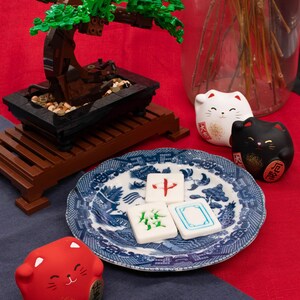 Op de afbeelding: Een miniatuur bonsai boom gemaakt van bruine en groene blokken staat op een houten standaard. Een blauw-wit bord bevat drie mahjongtegels. Drie gelukskatbeeldjes in wit, zwart en rood zijn ook aanwezig.