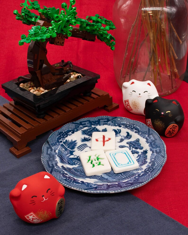 Op de afbeelding: Een miniatuur bonsai boom gemaakt van bruine en groene blokken staat op een houten standaard. Een blauw-wit bord bevat drie mahjongtegels. Drie gelukskatbeeldjes in wit, zwart en rood zijn ook aanwezig.