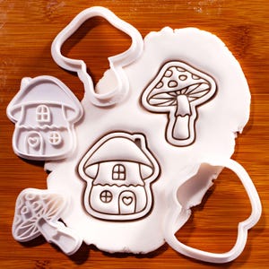 May include: A collection of white plastic cookie cutters in the shapes of mushrooms and mushroom-shaped houses, pressed into a flat piece of white dough. The cutters feature detailed outlines of windows, doors, and gills, creating impressions.