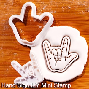 May include: White plastic cookie cutters in the shape of a hand sign and a heart. The hand sign is pressed into white dough, and the text "Hand Sign ILY Mini Stamp" is at the bottom.