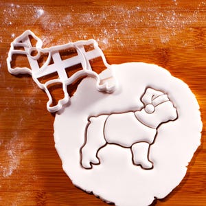 Op de afbeelding: Een witte plastic bulldog koekjessnijder naast een bulldogvormige koek op een houten oppervlak. De koek draagt een kerstmuts en een trui. Bloem is lichtjes rond de koek en de snijder gestrooid.