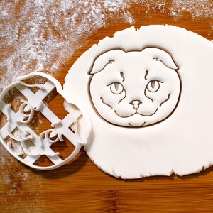 以下が含まれることがあります： 白い猫の形をしたクッキー型と、切り抜かれたクッキー。クッキー型は白いプラスチック製で、猫の顔の精巧なデザインが施されています。切り抜かれたクッキーも白く、猫の顔が描かれています。