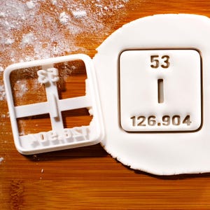 May include: White plastic cookie cutter shaped like a periodic table element. The element is iodine, with the atomic number 53 and atomic weight 126.904.