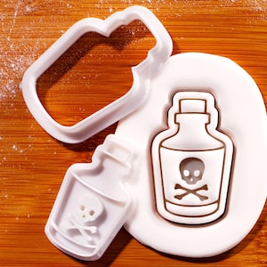 May include: A white plastic cookie cutter and a fondant design of a poison bottle with a skull and crossbones symbol. The cutter is in the shape of the bottle and has a handle. The fondant design is on a wooden surface.