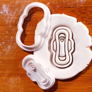 May include: White plastic cookie cutters in the shape of a sanitary pad. One cutter is a complete outline, another is a detailed impression, and the third is a smaller version. The cutters are on a wooden surface with a white dough.