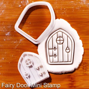May include: A white plastic cookie cutter and a matching stamp in the shape of an arched door with vertical panels, a circular window, and hinges. One piece is pressed into white dough, showing the embossed door design with the text Fairy Door Mini Stamp.