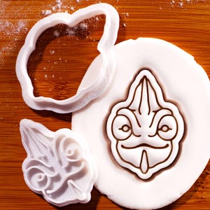 May include: Two white plastic cookie cutters and a circular piece of white dough. One cutter is an outline of a chameleon head, and the other is a detailed stamp featuring the facial features of a chameleon, pressed into the white dough.