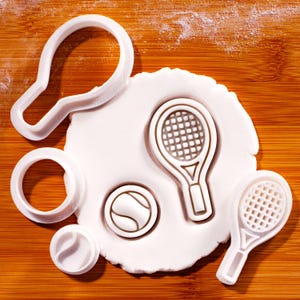 Könnte beinhalten: Weiße Ausstecher in Form von Tennisschlägern und einem Tennisball liegen auf einer Holzoberfläche. Daneben befindet sich ein weißer Teig mit Tennis-Schläger- und Ball-Abdrücken.