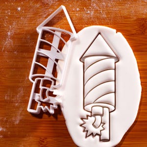 Könnte beinhalten: Weißer Ausstecher in Form einer Feuerwerksrakete, neben einer Feuerwerksraketenform, die in weißen Teig gedrückt wurde. Der Ausstecher hat ein detailliertes Design, einschließlich des Raketenkörpers, einer spitzen Spitze und eines Sterns am Boden.