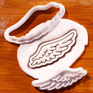 Peut inclure: Un emporte-pièce en forme d'aile d'ange blanc et un biscuit en forme d'aile sur une surface en bois. L'emporte-pièce est en plastique blanc et a un bord ondulé. Le biscuit en forme d'aile est blanc avec un contour marron.