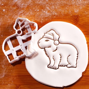 Op de afbeelding: Een witte capibara-koekjessnijder en een capibara-vormige koek met een kerstmuts op een houten oppervlak. De uitsteker ligt naast de koek, die in wit deeg is gedrukt. De koekjessnijder is gemaakt van wit plastic.