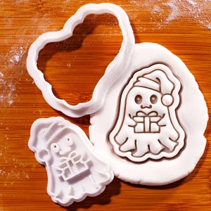 May include: White plastic cookie cutters in the shape of a ghost wearing a Santa hat and holding a gift. One cutter is a ghost shape, and the other is a heart shape. The ghost is pressed into white dough.