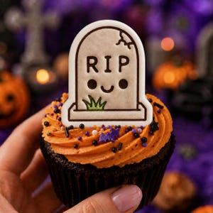 May include: A chocolate cupcake topped with orange frosting, multi-coloured sprinkles, and a flat, light-coloured tombstone-shaped topper. The topper features a cartoon face, a small patch of green grass, and the black text RIP at the top.