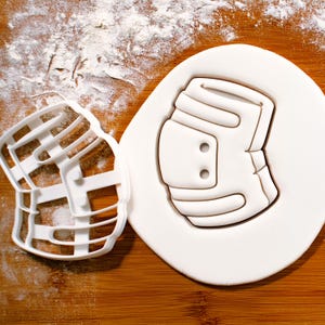 May include: A white plastic cookie cutter and a white cookie shaped like a hockey shoulder pad. The cookie cutter is on a wooden surface with flour. The cookie is on a white surface.