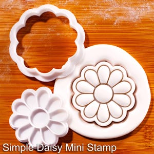 May include: White daisy-shaped cookie cutters and stamp on a wooden surface. The set includes a scalloped-edge cutter, a detailed stamp, and a smaller cutter. The text "Simple Daisy Mini Stamp" is visible at the bottom.