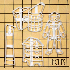 May include: Set of five white plastic cookie cutters shaped like a beekeeper, a beehive, a honeycomb, a smoker, and a beekeeper's ladder. The cutters are on a brown gridded surface with the word "INCHES" printed on the bottom right corner.