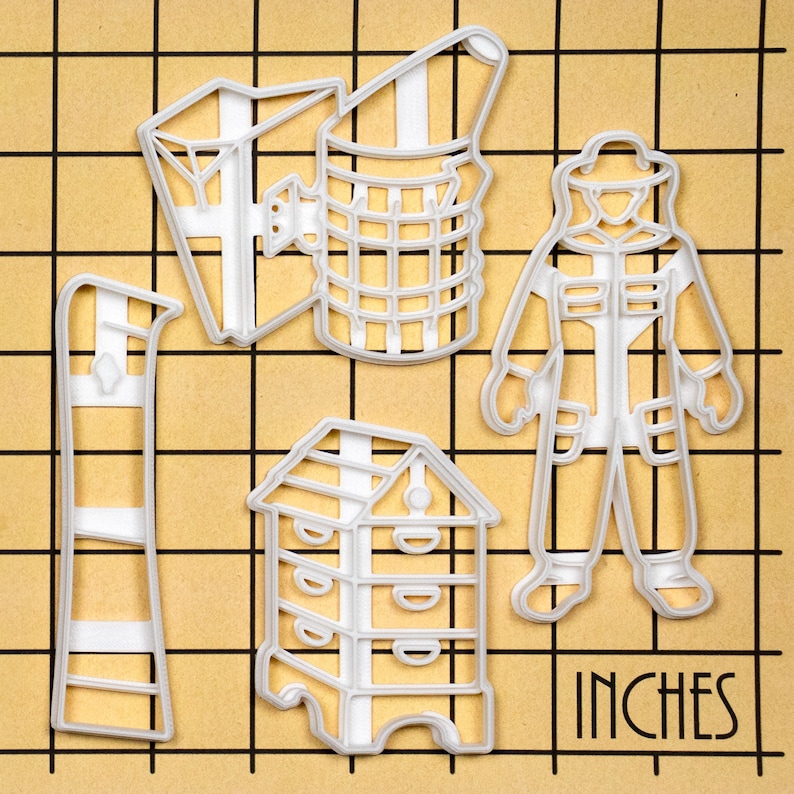 May include: Set of five white plastic cookie cutters shaped like a beekeeper, a beehive, a honeycomb, a smoker, and a beekeeper's ladder. The cutters are on a brown gridded surface with the word "INCHES" printed on the bottom right corner.