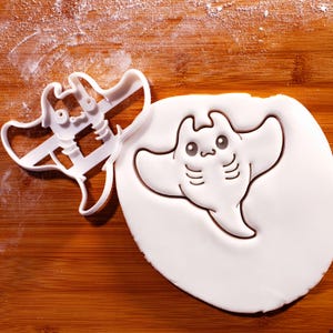 Peut inclure: Un emporte-pièce en plastique blanc en forme de raie manta de dessin animé, à côté d'un biscuit blanc avec le même motif. L'emporte-pièce et le biscuit sont sur une surface en bois avec de la farine.