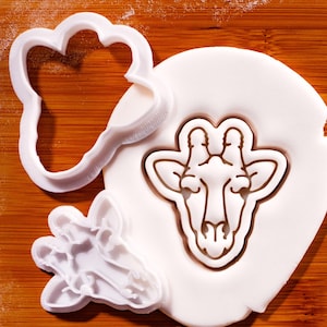 Könnte beinhalten: Eine weiße Keramik-Tasse mit schwarzem Henkel und dem Text "But First, Coffee" in fetter, schwarzer Schrift. Die Tasse steht vor einem verschwommenen Hintergrund, der eine gemütliche Umgebung suggeriert. Die Tasse ist etwa 10 cm hoch.