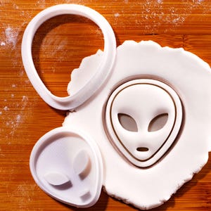 May include: White plastic alien-shaped cookie cutters and a circular cutter on a wooden surface. One cutter is pressed into white dough, creating an alien face impression. The other cutter is a complete alien face, and the third is a circular cutter.
