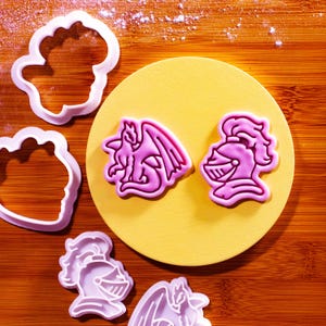 May include: Pink plastic cookie cutters shaped like a dragon, a knight's helmet, and a cloud. The cutters are on a yellow plate and a wooden surface.
