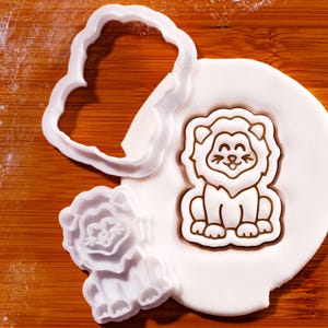 May include: A white plastic lion-shaped cookie cutter next to a lion-shaped cookie on a wooden surface. The cookie cutter is in the shape of a cloud. The cookie is brown and white.