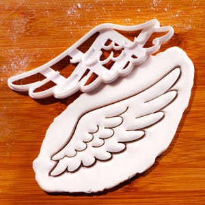 Peut inclure: Un emporte-pièce blanc en forme d'aile d'ange en plastique à côté d'un morceau de pâte en forme d'aile blanche. Le découpoir est posé sur la pâte, le tout sur une surface en bois.