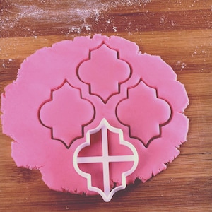 May include: A white plastic cookie cutter in the shape of a quatrefoil, also known as a four-leaf clover, is on top of pink fondant. Three other quatrefoil shapes have been cut out of the fondant.