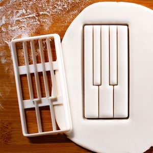 Op de afbeelding: Twee witte plastic koekjesvormen, een in de vorm van een pianoklavier en de andere met een rasterpatroon. De vormen liggen op een houten oppervlak met wit poeder.