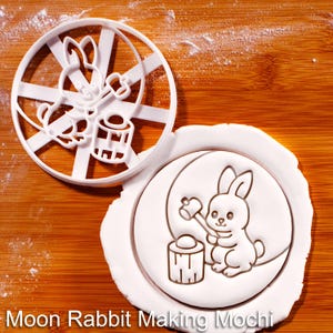 May include: White plastic cookie cutter with a rabbit design, next to a stamped cookie. The design features a rabbit holding a mallet and a mochi cake. The text "Moon Rabbit Making Mochi" is at the bottom.