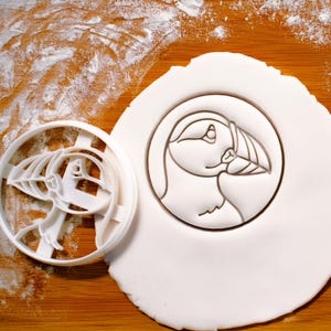 May include: A white plastic cookie cutter and a puffin-shaped imprint on white dough. The cutter is a circular shape with the puffin design cut out. The puffin imprint is in a circle, with a detailed outline of the bird's head and beak.