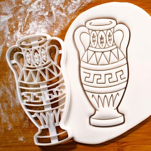 May include: White plastic cookie cutter in the shape of a Greek amphora or vase with a geometric design. The cutter is on a white surface with flour.