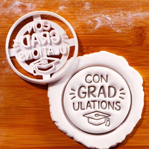 May include: White plastic cookie cutter with the words "CON GRAD ULATIONS" and a graduation cap design. The cutter is on a wooden surface with a cut-out cookie.