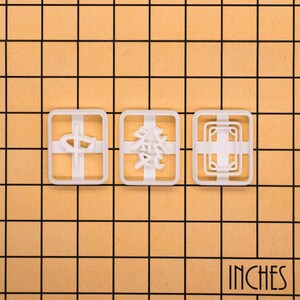 Op de afbeelding: Drie witte plastic koekjessnijders met verschillende ontwerpen: een kruis, Chinese karakters en een geometrisch patroon. De snijders zijn rechthoekig met afgeronde hoeken. Het woord "INCHES" staat rechtsonder gedrukt.