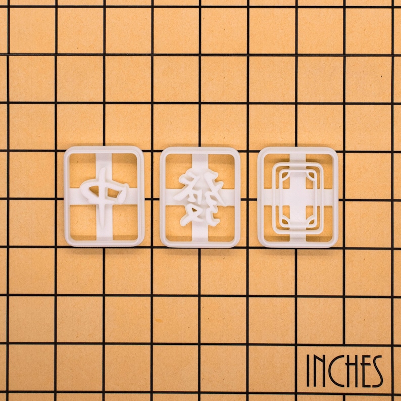 Op de afbeelding: Drie witte plastic koekjessnijders met verschillende ontwerpen: een kruis, Chinese karakters en een geometrisch patroon. De snijders zijn rechthoekig met afgeronde hoeken. Het woord "INCHES" staat rechtsonder gedrukt.