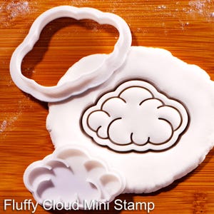 Peut inclure: Un emporte-pièce en plastique blanc en forme de nuage et un tampon assorti en forme de nuage. Le tampon est pressé dans une pâte blanche, créant un contour de nuage détaillé. Le texte Fluffy Cloud Mini Stamp est visible en bas.