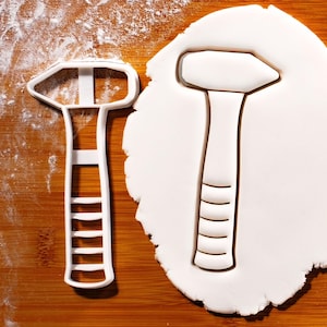 May include: A white plastic cookie cutter shaped like a hammer. The cutter is on a wooden surface with a white background.