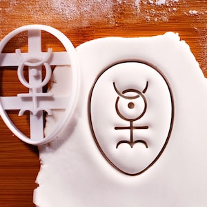 May include: A white plastic cookie cutter with a unique symbol, next to a white cookie with the same embossed design. The symbol features curved lines, circles, and a horizontal line with three downward curves. The background is a wooden surface.