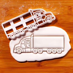 May include: A white plastic cookie cutter in the shape of a semi-truck, placed on a wooden surface. The cutter is next to a semi-truck shaped cookie. The cookie cutter has a detailed design of the truck's cab, wheels, and trailer.