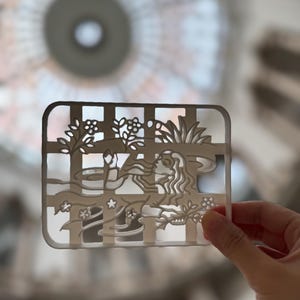May include: A rectangular, off-white decorative piece with a grid pattern and a cut-out design featuring a woman's profile, flowers, and foliage. The piece is held up against a blurred background.
