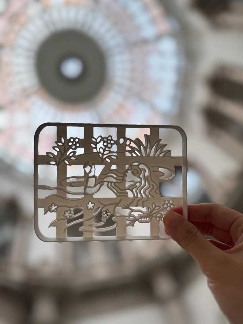 May include: A rectangular, off-white decorative piece with a grid pattern and a cut-out design featuring a woman's profile, flowers, and foliage. The piece is held up against a blurred background.