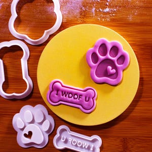 May include: A collection of pink and white dog-themed cookie cutters and stamps. The stamps include a paw print with a heart and a bone-shaped stamp with the text "I WOOF U". The items are arranged on a yellow circle and a wooden surface.
