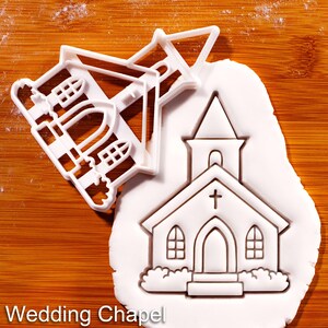 May include: A white plastic cookie cutter shaped like a church with a steeple and a cross, positioned next to a piece of white rolled dough that has been imprinted with the same church design, featuring arched windows and a central doorway.