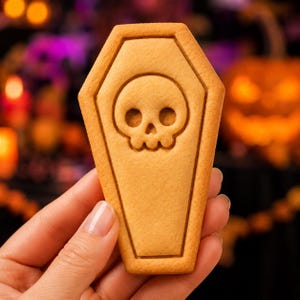 May include: A light brown, coffin-shaped biscuit featuring a debossed skull design in the centre. The biscuit has a smooth, baked texture with a distinct outline around the perimeter and a detailed skull motif pressed into the surface of the dough.